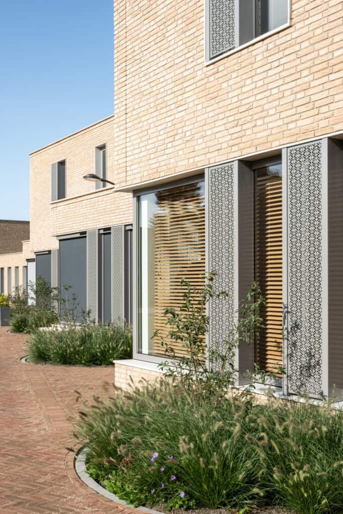 Kleurr verpleegkamergordijnen en houten jaloezieën aan een modern herenhuis, gevelbekleding met metalen decoraties en een groene tuin, ontworpen door Studio Redd voor een stijlvolle en functionele buitenruimte.