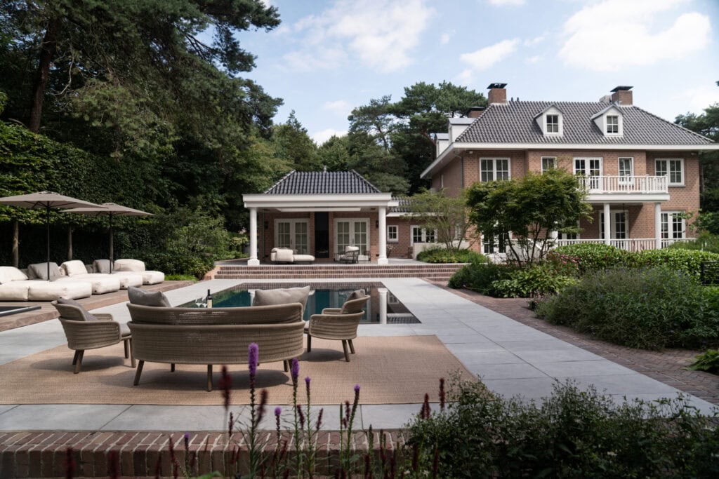 Luxe moderne tuin met uitnodigend zwembad, rieten loungestoelen en comfortabele zitbanken, omgeven door groene tuinplanten en hoge bomen, bij een rietgedekte villa in een bosrijke omgeving.