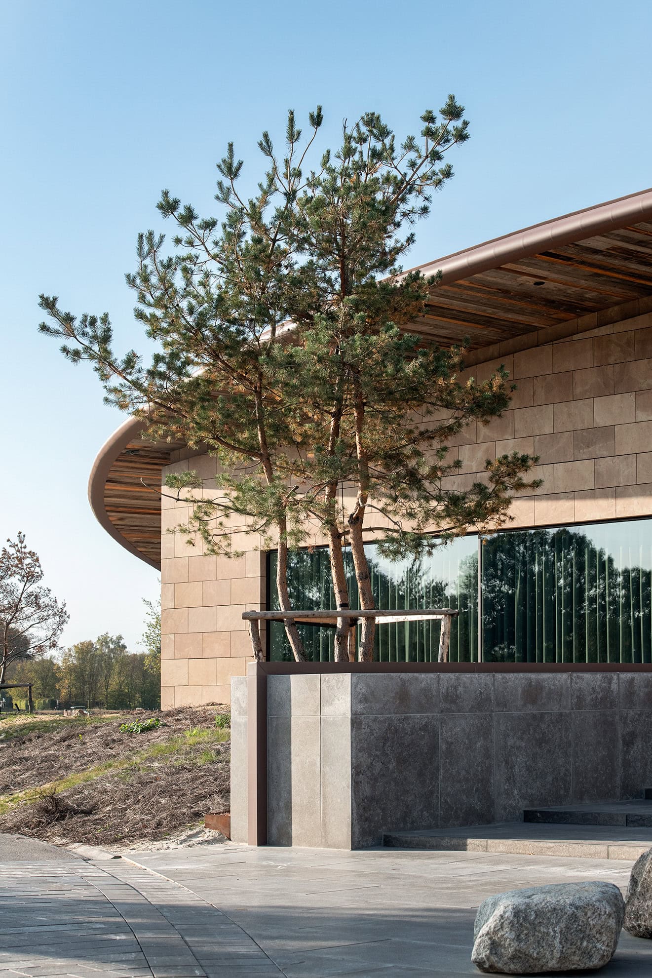 Moderne architectuur met natuurlijke elementen, zoals een houten omheining en een grote raam, gecombineerd met een strak betegeld buitengevel en een jonge dennenboom, ontworpen door Studio REDD.
