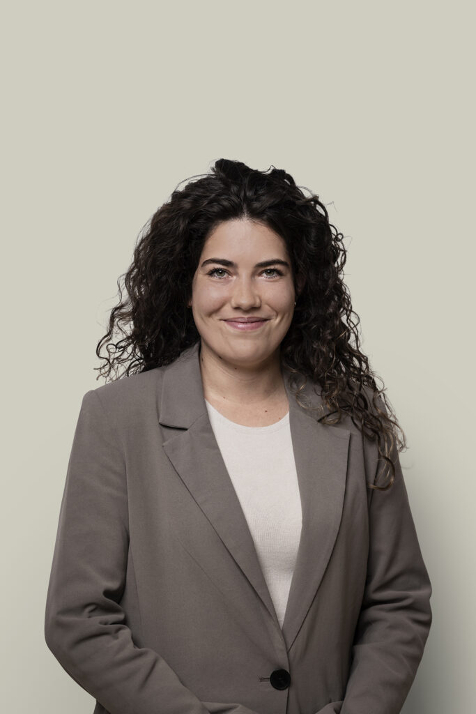 Strakke zakelijke portretfoto van een vrouw met donkere, krullende haar en een vriendelijke glimlach, professioneel voor bedrijf, LinkedIn, en marketingdoeleinden.