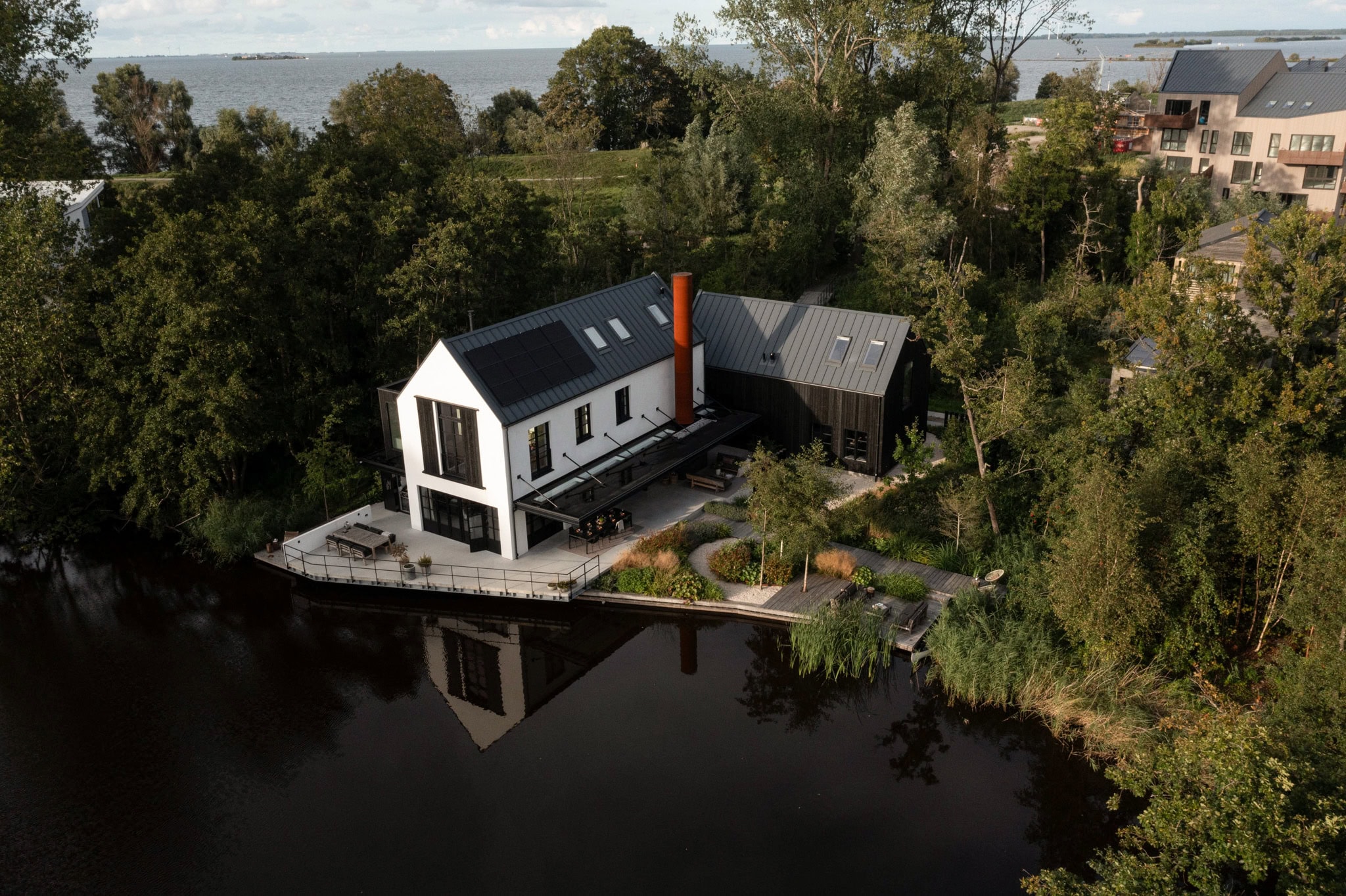 Luchtfoto van een modern huis met zwarte en witte gevels, gelegen aan een rustige rivier omringd door groen bos en bomen, met een terras en tuin.