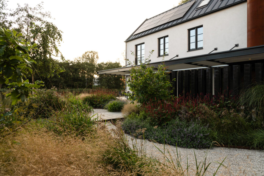 Moderne tuin met een duurzame woning, omgeven door diverse planten, struiken en een grindpad, met zonnepanelen op het dak, passend bij het thema 'Automatische concepten'.