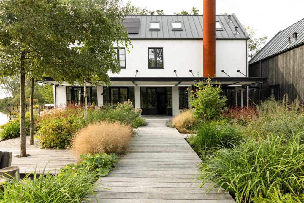 Moderne woning met groene tuin en houten wandelpad, omgeven door diverse planten en bomen, onder een heldere hemel.