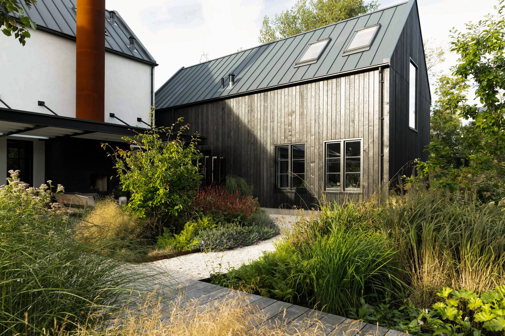 Modern houten huis met tuin en wandelpad, omgeven door groen en natuurlijke beplanting, onder een heldere hemel.