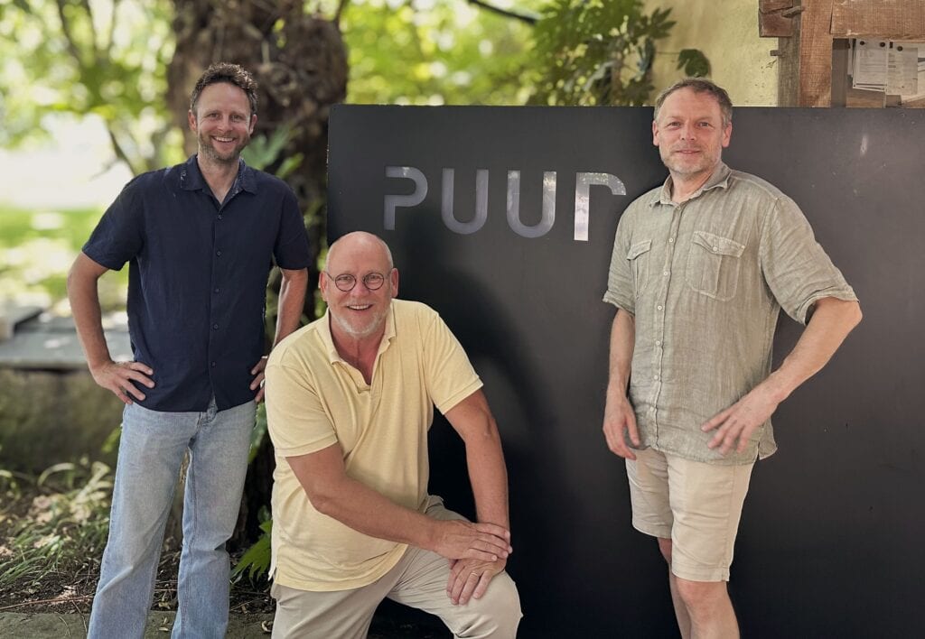 Drie mannen bij Studio REDD voor een zwart bord met de tekst PUUR, in een groene buitenomgeving, symboliseert de overname van PUUR groenprojecten.