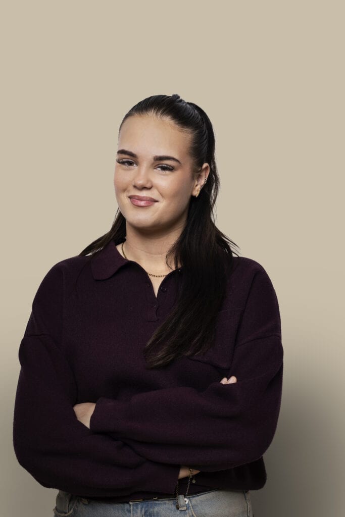 Portret van jonge vrouw met donkere haar en casual kleding, lachend en zelfverzekerd, op neutrale achtergrond.