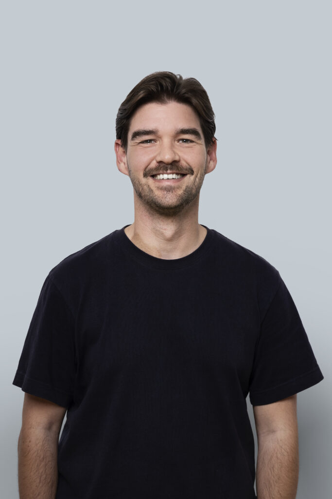 Portret van een lachende man met donkere haar en een zwarte T-shirt, voor een lichte achtergrond, geschikt voor bedrijfs- en portretfotografie.