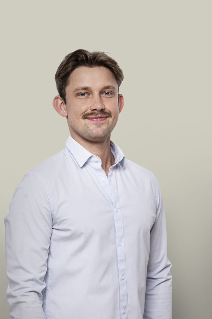Man met lichte haarkleur en lichte ogen, glimlachend in wit overhemd, professionele uitstraling, neutrale achtergrond, geschikt voor zakelijke of professionele context.